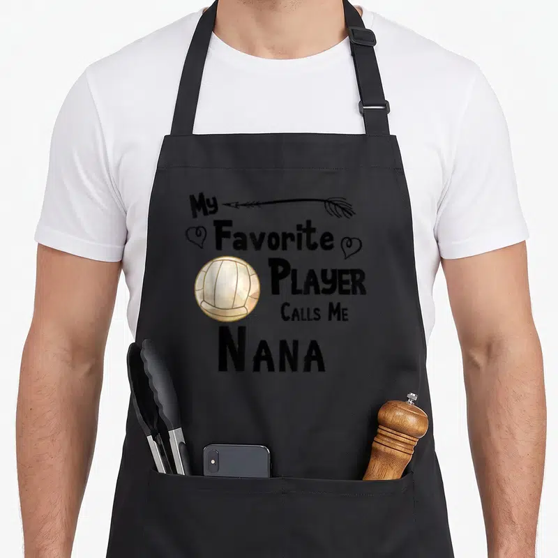 Game nana aprons - Stacked text My Favorite Player Calls Me Nana with arrow, two hearts, and cream volleyball — shown on a black cotton canvas apron against a white background