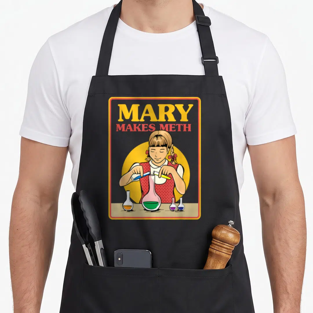 Mary sarcastic apron - Cartoon girl pouring blue and yellow liquids into a flask with green contents under MARY MAKES — shown on a black cotton canvas apron against a white background