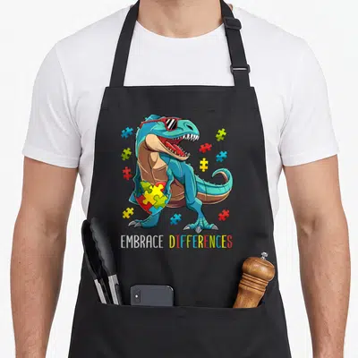 Autism dinosaur apron - Blue cartoon dinosaur with red sunglasses holding a multicolor puzzle heart with scattered — shown on a black cotton canvas apron against a white background