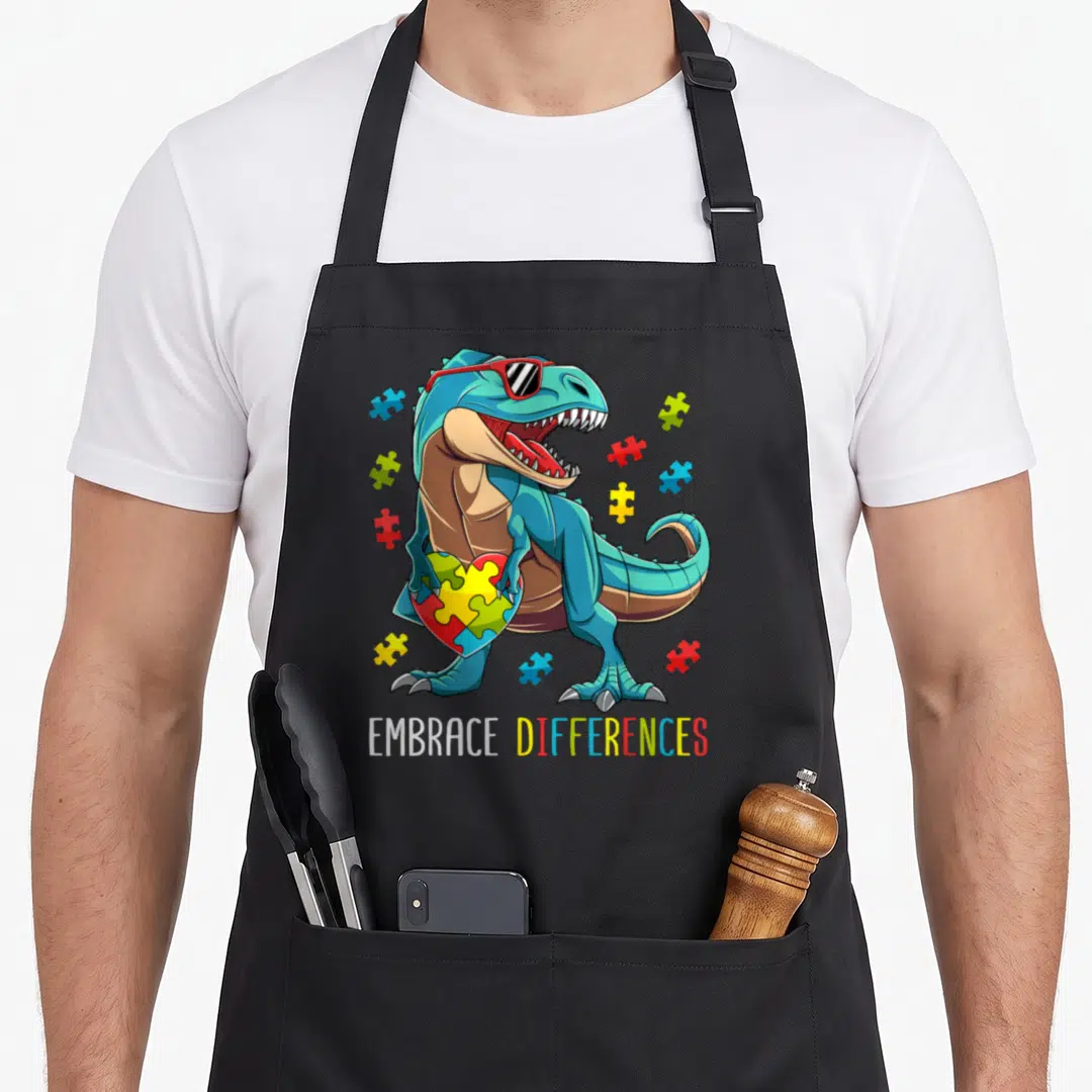 Autism dinosaur apron - Blue cartoon dinosaur with red sunglasses holding a multicolor puzzle heart with scattered — shown on a black cotton canvas apron against a white background