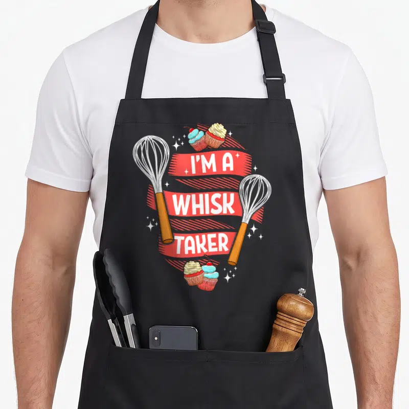 Pun baker apron - Two whisks flank red banner with I'M A WHISK TAKER text and cupcakes — shown on a black cotton canvas apron against a white background