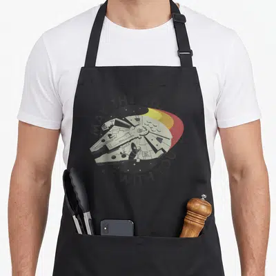 May falcon apron - Retro Millennium Falcon in starry circle with yellow and red streaks and arched text — shown on a black cotton canvas apron against a white background