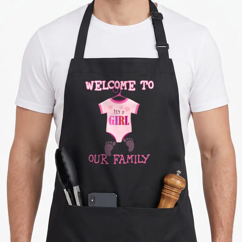 Shower family aprons - Pink onesie with It's a GIRL text, purple footprints, and WELCOME TO OUR FAMILY lettering — shown on a black cotton canvas apron against a white background