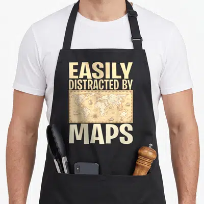 Geography teacher apron - EASILY DISTRACTED BY MAPS text above and below a beige vintage world map with ships — shown on a black cotton canvas apron against a white background
