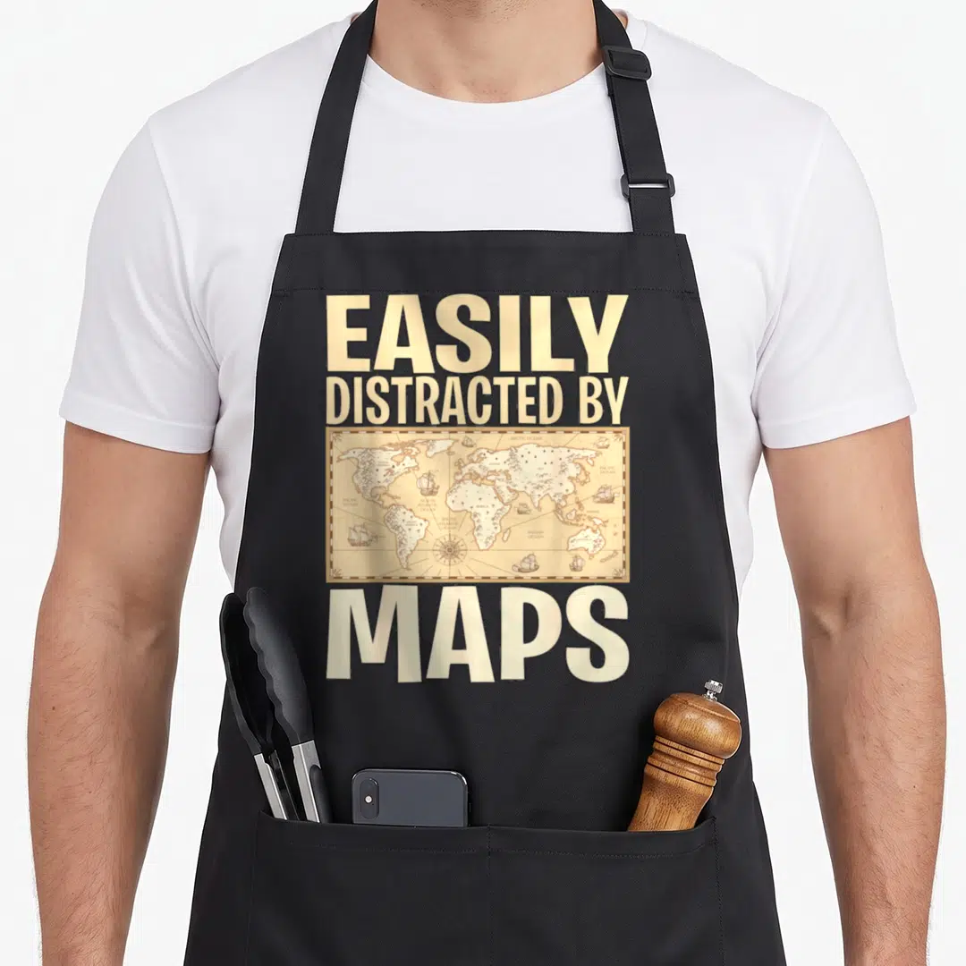 Geography teacher apron - EASILY DISTRACTED BY MAPS text above and below a beige vintage world map with ships — shown on a black cotton canvas apron against a white background
