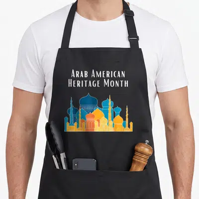 Arab heritage aprons - Stacked white text above a blue orange and yellow mosque skyline silhouette — shown on a black cotton canvas apron against a white background