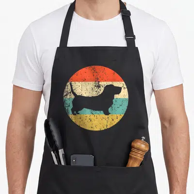 Hound dog apron - Grey basset hound silhouette on red cream teal yellow striped circle — shown on a black cotton canvas apron against a white background