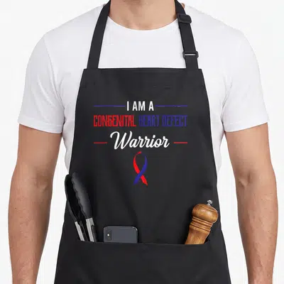 Congenital ribbon apron - Stacked text I AM A CONGENITAL HEART DEFECT Warrior with red and blue ribbon below — shown on a black cotton canvas apron against a white background