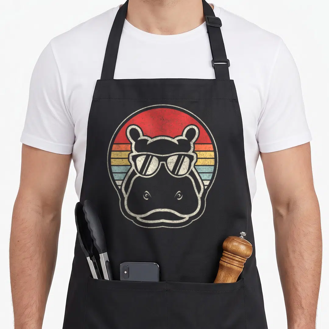 Sunglass hippo apron - Grey hippo head with sunglasses in front of red orange yellow and blue striped circle — shown on a black cotton canvas apron against a white background