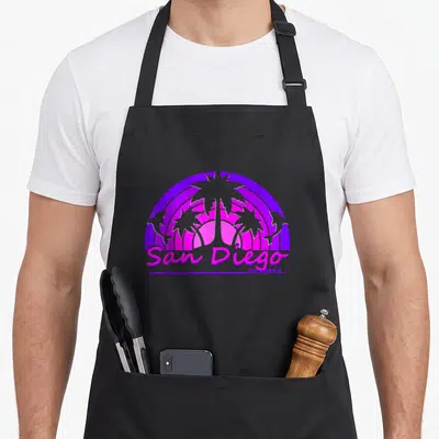 San charlottesville apron - Retro purple and pink sun graphic with palm trees, two dolphins, and San Diego California — shown on a black cotton canvas apron against a white background