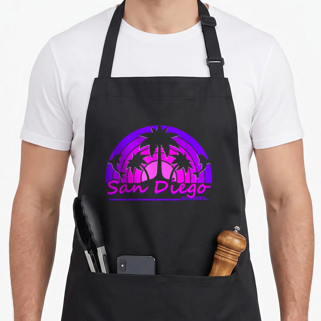 San charlottesville apron - Retro purple and pink sun graphic with palm trees, two dolphins, and San Diego California — shown on a black cotton canvas apron against a white background