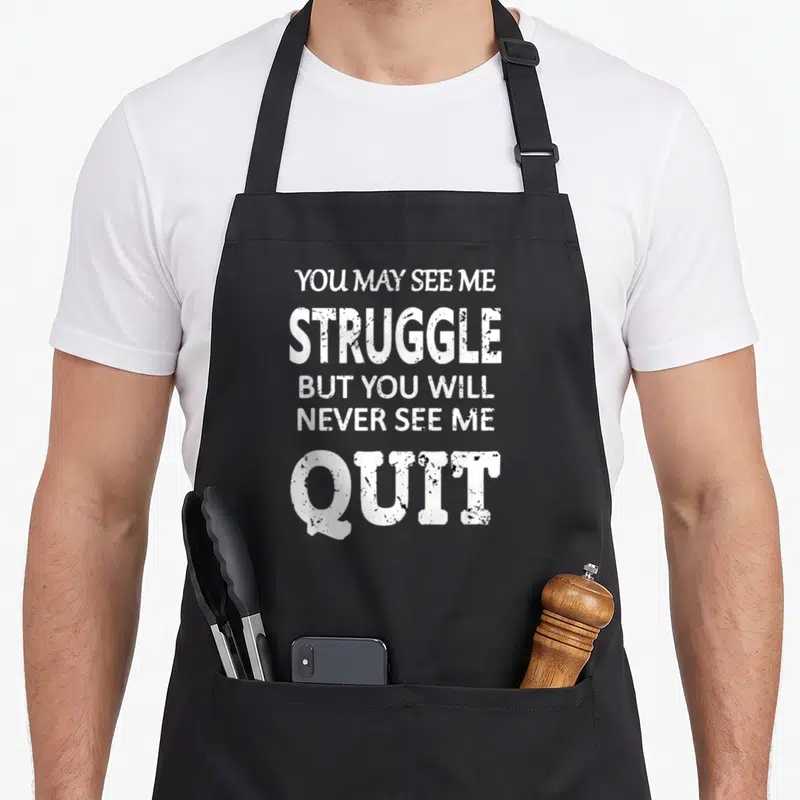 Inspirational wedding apron - White stacked text reading YOU MAY SEE ME STRUGGLE BUT YOU WILL NEVER SEE ME QUIT — shown on a black cotton canvas apron against a white background