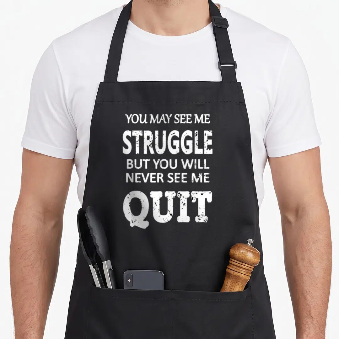 Inspirational wedding apron - White stacked text reading YOU MAY SEE ME STRUGGLE BUT YOU WILL NEVER SEE ME QUIT — shown on a black cotton canvas apron against a white background