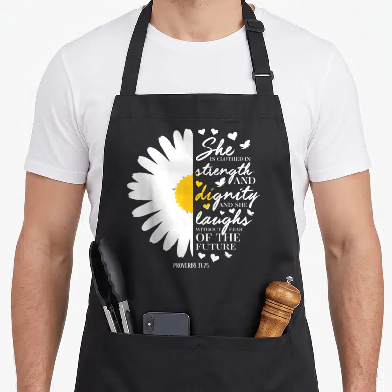 Daisy bible verse apron - Half daisy beside She is clothed in strength and dignity text with hearts and butterflies — shown on a black cotton canvas apron against a white background