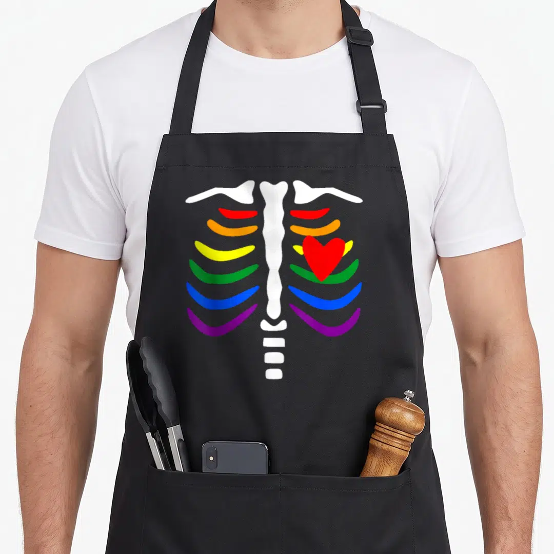 Skeleton gay pride apron - White ribcage with rainbow colored ribs and red heart graphic — shown on a black cotton canvas apron against a white background