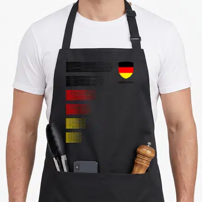 Germany patriotic apron - Shield with German flag colors and GERMANY text beside six colored horizontal line groups — shown on a black cotton canvas apron against a white background