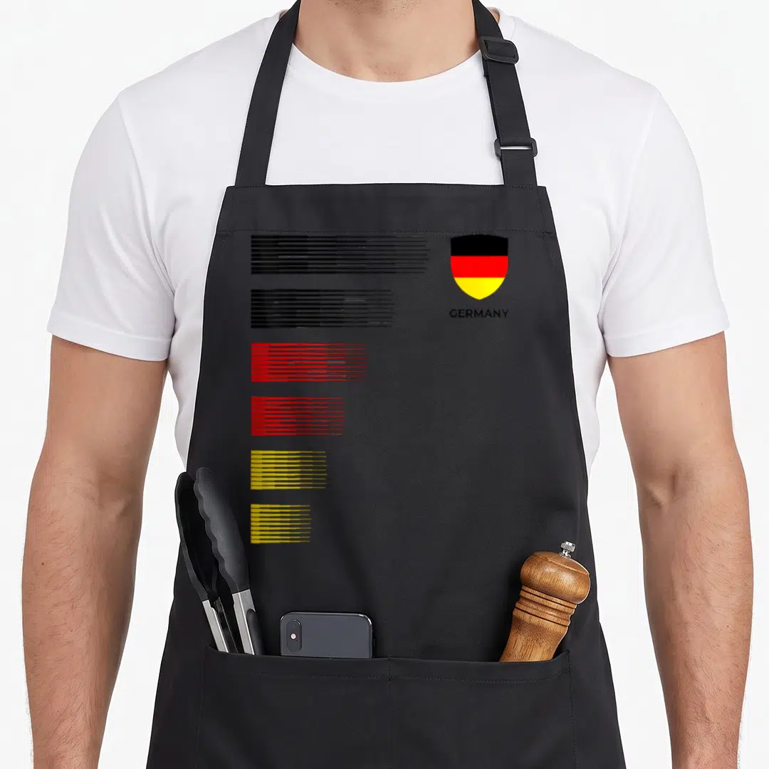 Germany patriotic apron - Shield with German flag colors and GERMANY text beside six colored horizontal line groups — shown on a black cotton canvas apron against a white background