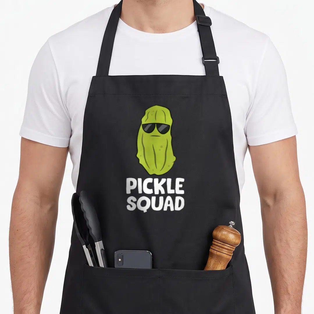 Dill food aprons - Green cartoon pickle with sunglasses above white PICKLE SQUAD text — shown on a black cotton canvas apron against a white background