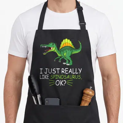 Spinosaurus dinosaur apron for men - Cartoon green spinosaurus with orange sail above text I Just Really Like — shown on a black cotton canvas apron against a white background