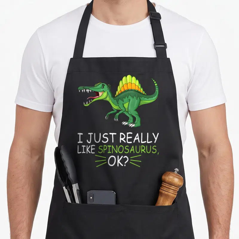 Spinosaurus dinosaur apron for men - Cartoon green spinosaurus with orange sail above text I Just Really Like — shown on a black cotton canvas apron against a white background