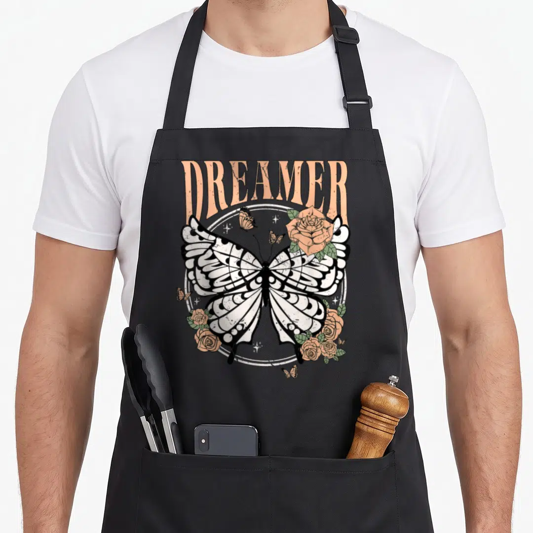 Dreamer butterfly apron - DREAMER text above large butterfly wings surrounded by roses and stars — shown on a black cotton canvas apron against a white background