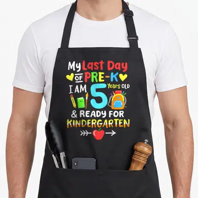 Pre another year older apron - My Last Day of Pre-K text with number 5, backpack, pencils, and apple in cartoon style — shown on a black cotton canvas apron against a white background