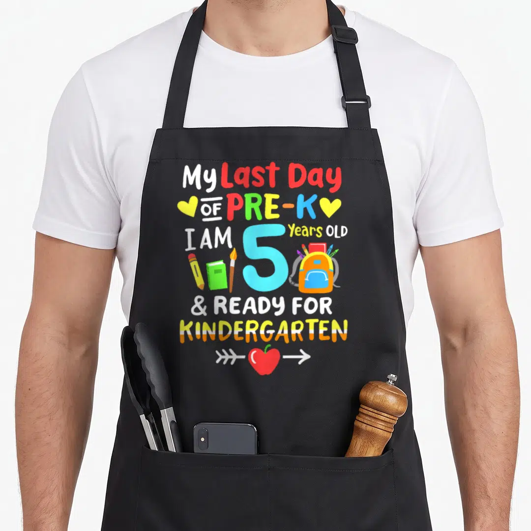 Pre another year older apron - My Last Day of Pre-K text with number 5, backpack, pencils, and apple in cartoon style — shown on a black cotton canvas apron against a white background