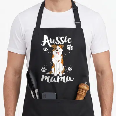 Mama australian aprons - Cartoon dog with white brown orange fur and four paw prints under Aussie mama text — shown on a black cotton canvas apron against a white background