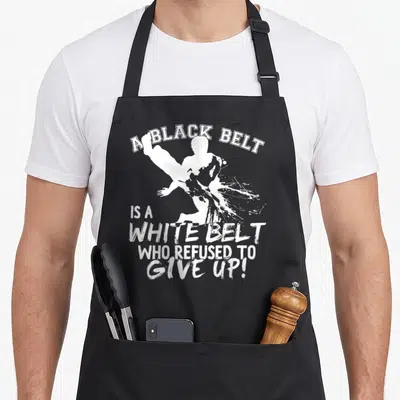 Belt martial apron - White distressed text A Black Belt Is A White Belt Who Refused To Give Up above a kicking martial — shown on a black cotton canvas apron against a white background