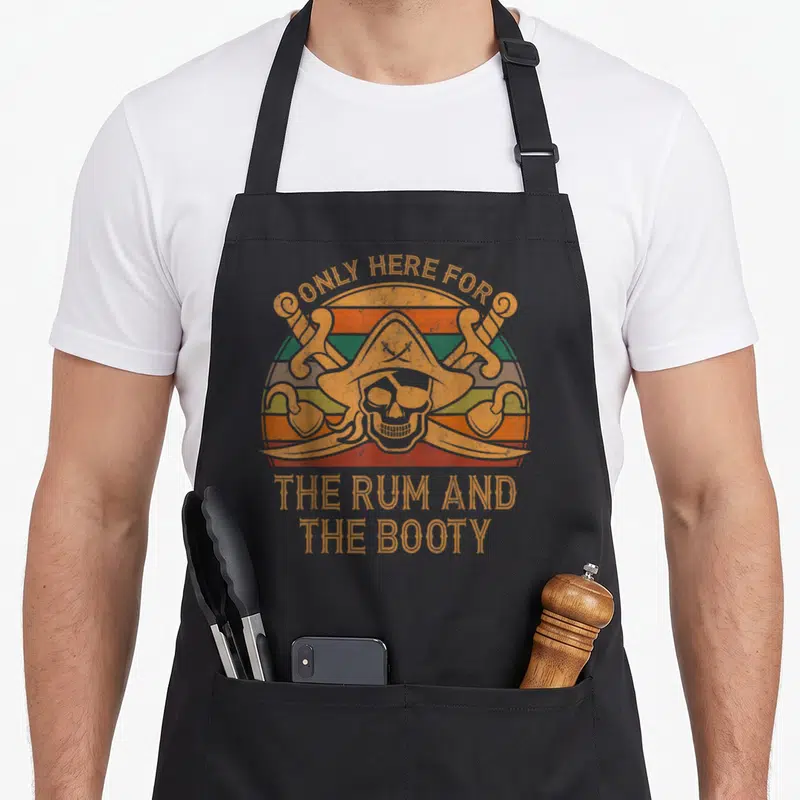 Rum halloween apron - Skull in pirate hat with crossed cutlasses and hooks over striped sun, text ONLY HERE FOR THE RUM — shown on a black cotton canvas apron against a white background