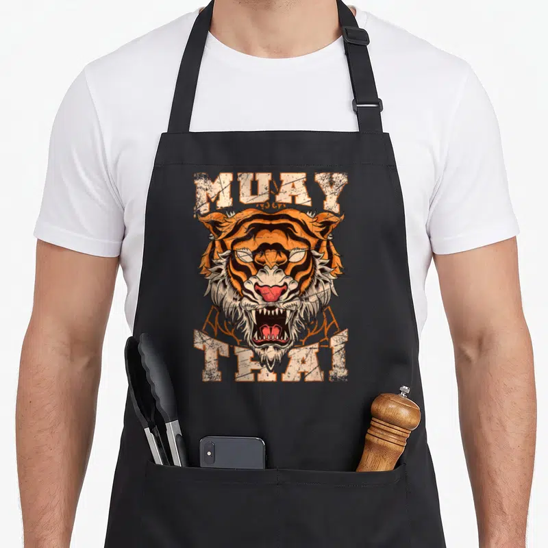 Asia thai girl apron - Distressed tiger head between MUAY and THAI text in orange and black — shown on a black cotton canvas apron against a white background