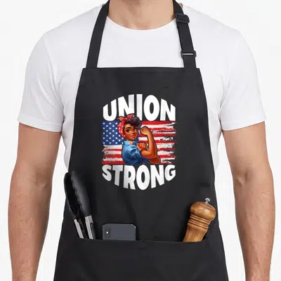 Union american flag apron - Union Strong text framing a flexing woman over a distressed US flag — shown on a black cotton canvas apron against a white background