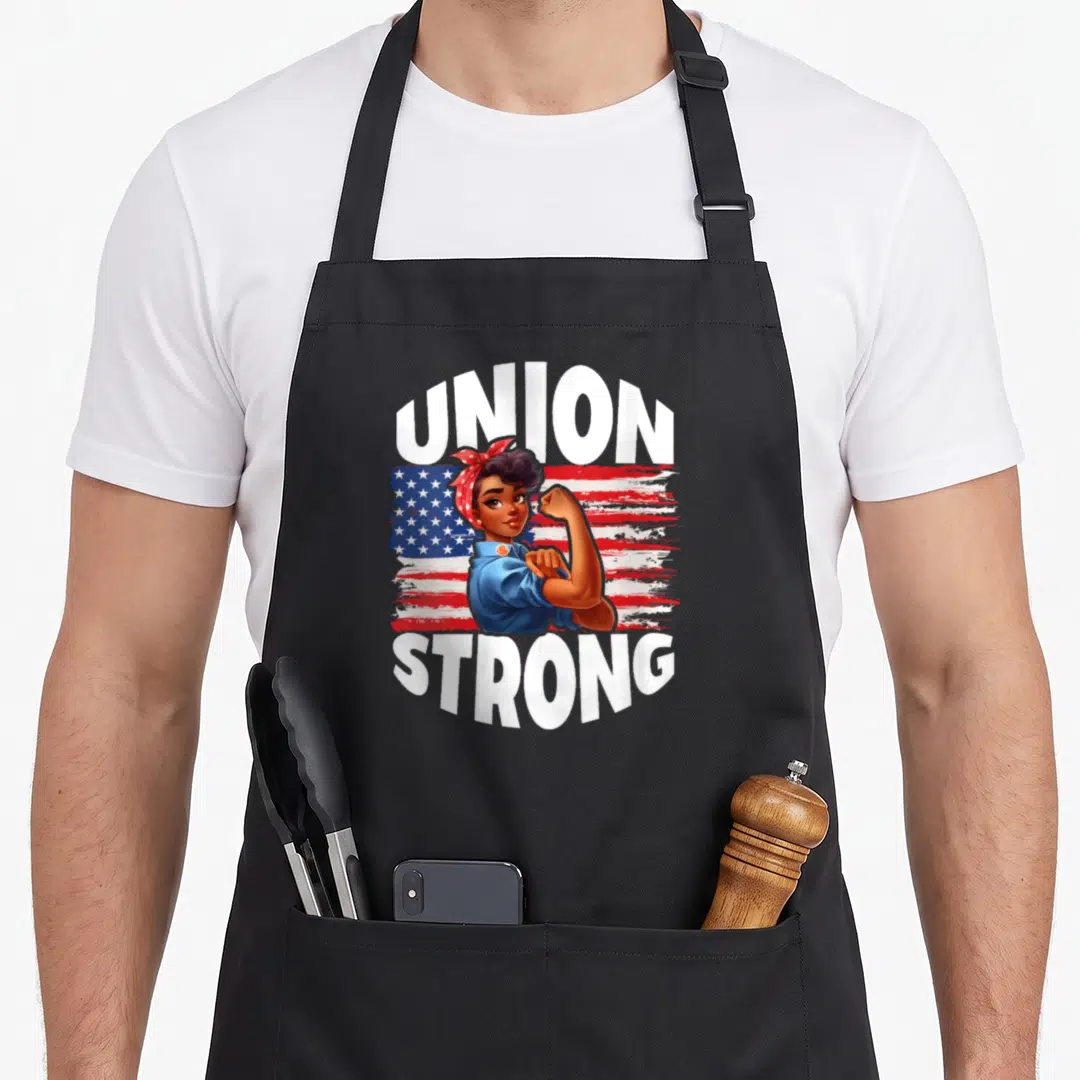 Union american flag apron - Union Strong text framing a flexing woman over a distressed US flag — shown on a black cotton canvas apron against a white background