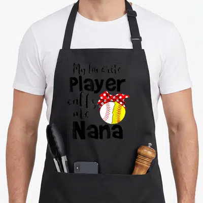 Player nana aprons - Stacked text next to a split yellow and white softball topped with a red polka dot bow — shown on a black cotton canvas apron against a white background