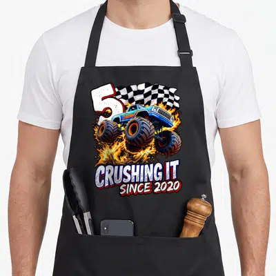 Monster 5th truck apron - Blue monster truck jumping through fire next to large number 5 and checkered flag — shown on a black cotton canvas apron against a white background