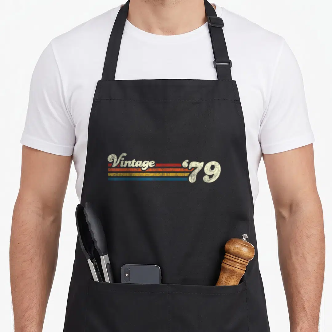 Casual birthday apron - Retro typography reading Vintage '79 with orange yellow and blue horizontal stripes — shown on a black cotton canvas apron against a white background