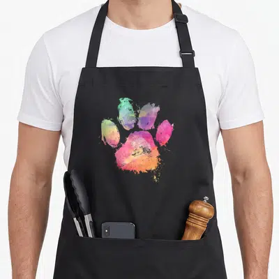 Watercolor paw print apron - Rainbow watercolor paw print in pink, orange, yellow, green, blue, and purple — shown on a black cotton canvas apron against a white background