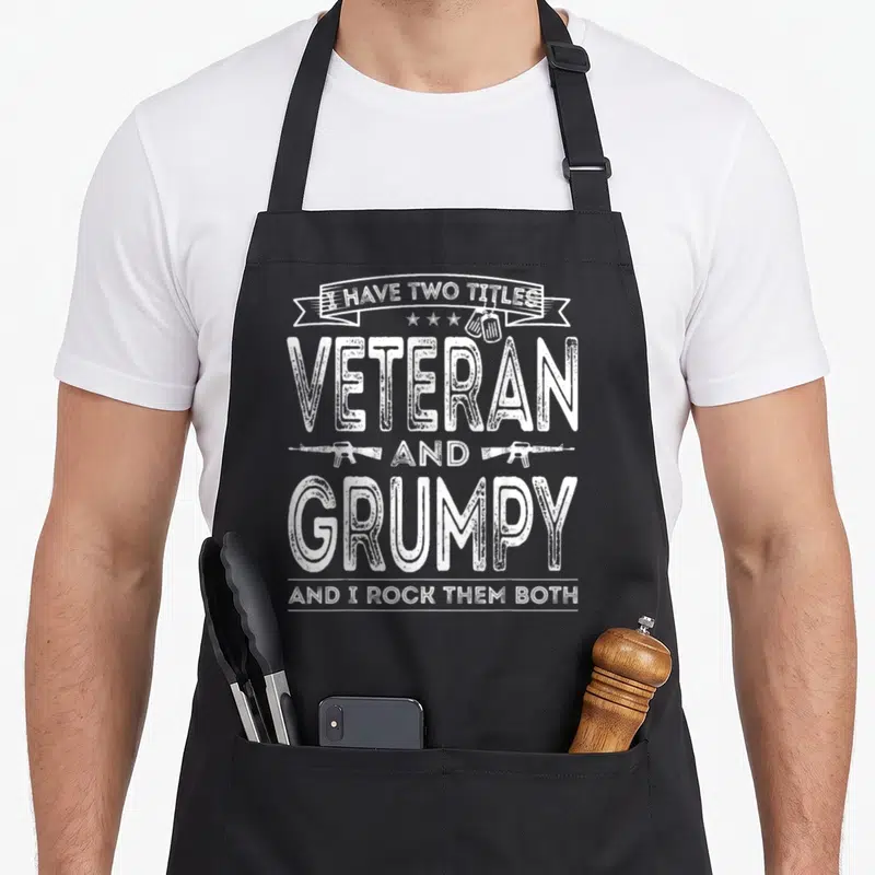 Title us aprons - White distressed text reading I HAVE TWO TITLES VETERAN GRUMPY with dog tags and rifles — shown on a black cotton canvas apron against a white background