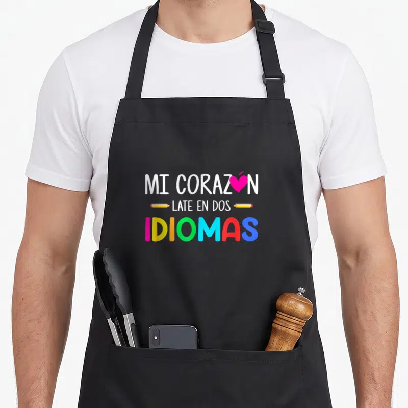 Corazon spanish aprons - MI CORAZON LATE EN DOS IDIOMAS text with pink heart and yellow pencils — shown on a black cotton canvas apron against a white background