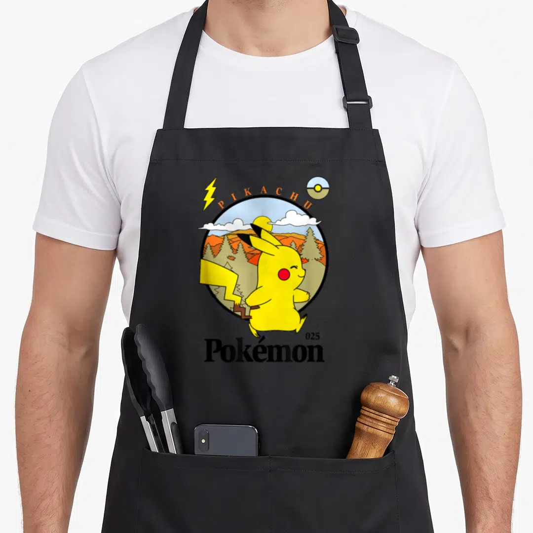 Mon outdoor apron - Pikachu yellow cartoon character inside circular mountain landscape with PIKACHU text and pokeball — shown on a black cotton canvas apron against a white background