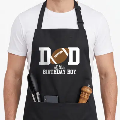 Funny football birthday apron - Dad of the Birthday Boy text with brown football graphic — shown on a black cotton canvas apron against a white background