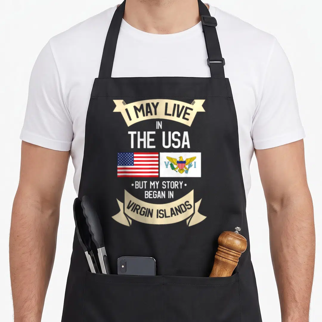 Root kitchen island apron - Stacked text about Virgin Islands roots flanking two flags in red white and blue — shown on a black cotton canvas apron against a white background