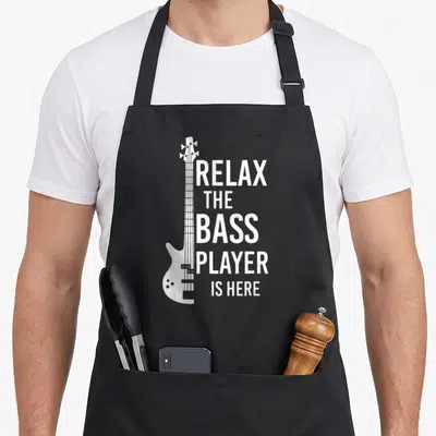 Relax player bass apron - White bass guitar graphic beside five lines of text reading Relax The Bass Player Is Here — shown on a black cotton canvas apron against a white background