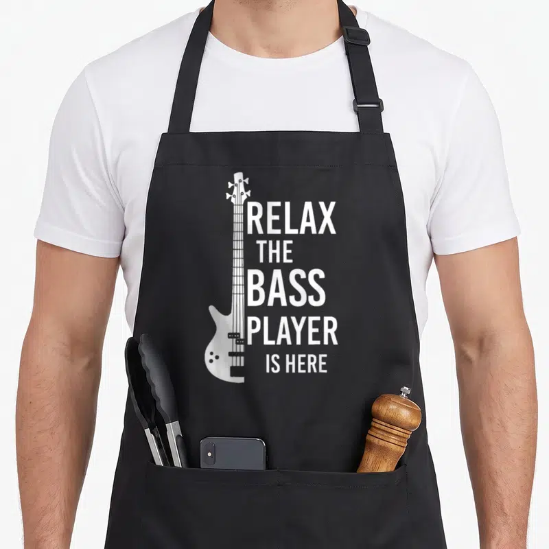 Relax player bass apron - White bass guitar graphic beside five lines of text reading Relax The Bass Player Is Here — shown on a black cotton canvas apron against a white background