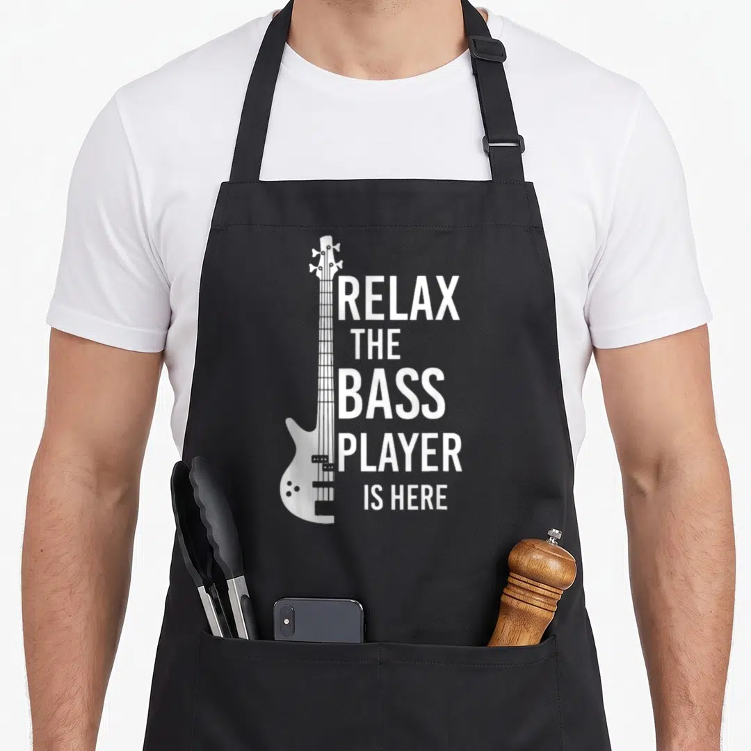 Relax player bass apron - White bass guitar graphic beside five lines of text reading Relax The Bass Player Is Here — shown on a black cotton canvas apron against a white background