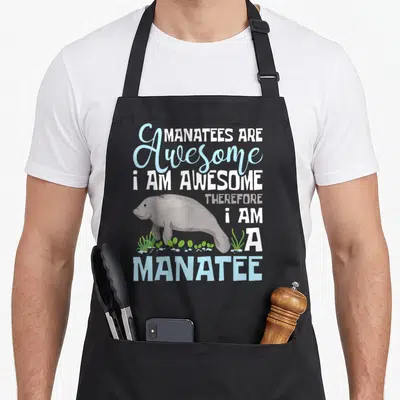 Zookeeper manatee apron - Cartoon manatee swimming among green seaweed and bubbles below stacked text reading MANATEES — shown on a black cotton canvas apron against a white background