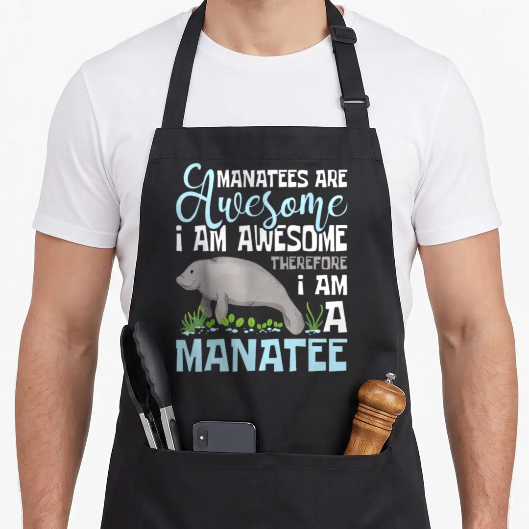 Zookeeper manatee apron - Cartoon manatee swimming among green seaweed and bubbles below stacked text reading MANATEES — shown on a black cotton canvas apron against a white background
