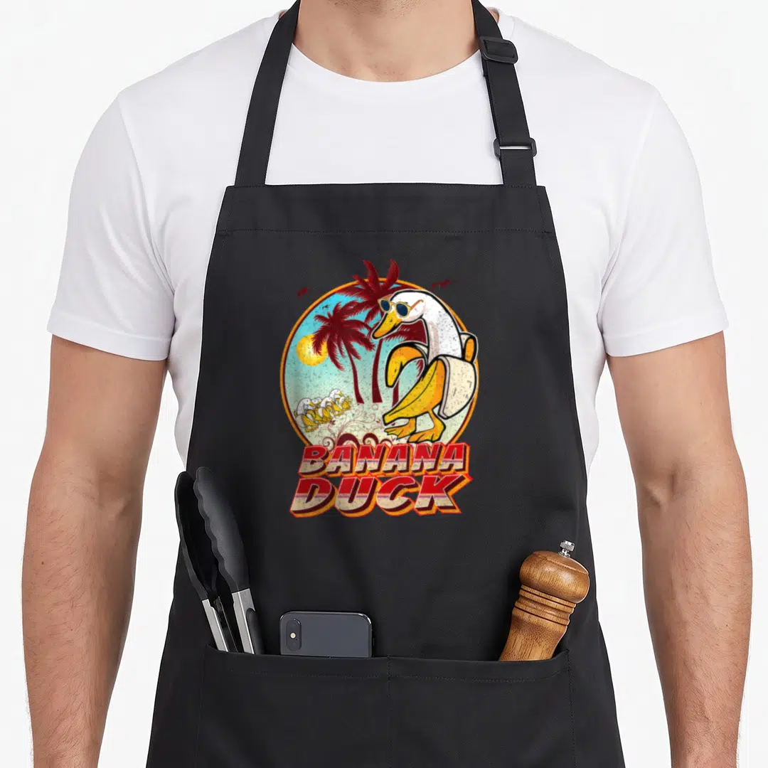 Banana duck apron - Retro duck in sunglasses holding banana peel in tropical circle with red stacked text — shown on a black cotton canvas apron against a white background