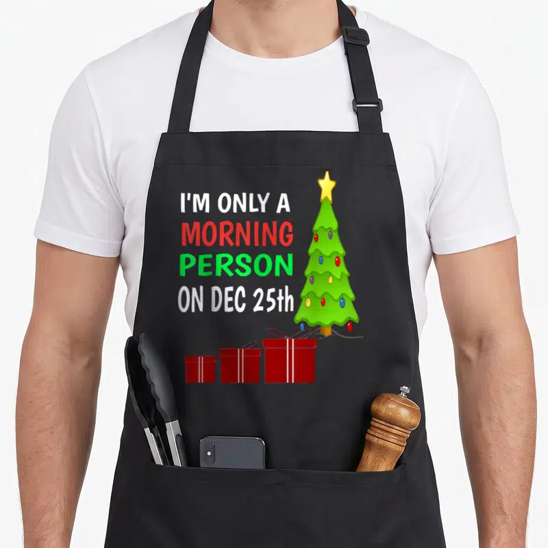 Merry women's christmas apron - I'M ONLY A MORNING PERSON ON DEC 25th text with green tree and three gifts — shown on a black cotton canvas apron against a white background