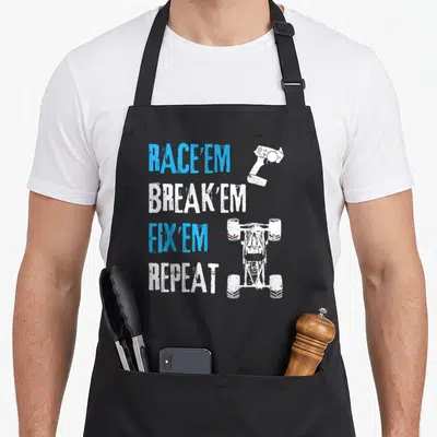 Remote car apron - Distressed blue and white text RACE'EM BREAK'EM FIX'EM REPEAT with RC car and controller silhouettes — shown on a black cotton canvas apron against a white background